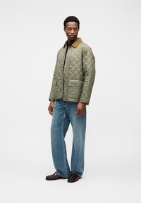 Καπιτονέ χακί μπουφάν με καφέ κορδούρι γιακά, μπροστινές τσέπες και κουμπιά, συνδυασμένο με ανοιχτό μπλε τζιν παντελόνι με φαρδιά πόδια και σκούρα παπούτσια.