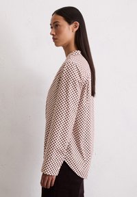 Chemise à manches longues de couleur rose clair avec un motif géométrique marron, présentant une coupe décontractée et un ourlet arrondi, fabriquée à partir d'un tissu texturé.