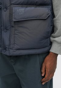 Chaleco acolchado azul marino con un bolsillo y detalle de logo, con una textura suave y diseño redondeado, usado sobre un suéter gris.