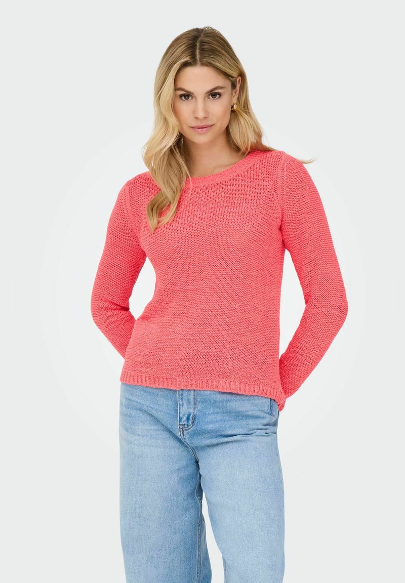 Femme aux cheveux longs blonds portant un pull en maille corail et un jean bleu clair, debout les mains derrière le dos sur un fond uni.