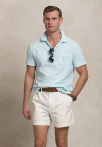 Lichtblauwe polo met korte mouwen en een zak, gecombineerd met witte shorts en een bruine gevlochten riem, geaccessoreerd met donkere zonnebril en een horloge.