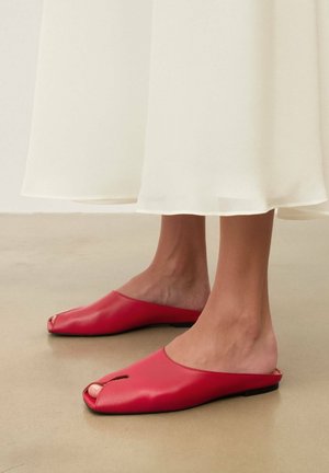 Pėdos, avinčios raudonus atvirais pirštais batus be užsegimo, derinamus su ilga balta suknele, stovinčios ant lygaus smėlio spalvos grindinio.