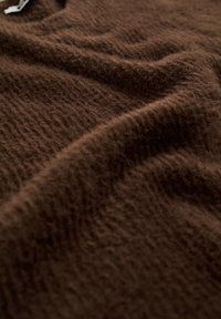 Brun texturerad tyg med en mjuk, fluffig yta, som har subtila horisontella linjer och mjuka veck, vilket indikerar ett lätt stickat material.