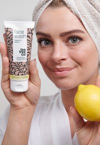 Gezichtspoeling tube met bloemmotief in roze en wit, gelabeld "tea tree olie," vastgehouden in één hand, gele citroen in de andere, tegen een lichte achtergrond.