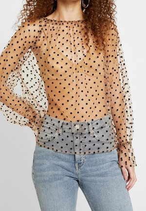 Kobieta w przezroczystej beżowej bluzce z długim rękawem w czarne kropki, na beżowym staniku, połączonej z jasnoniebieskimi dżinsami.