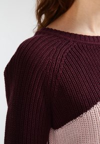 Nahaufnahme einer Person, die einen gerippten Strickpullover mit dunkelvioletten und hellrosanen diagonalen Farbstreifen trägt.
