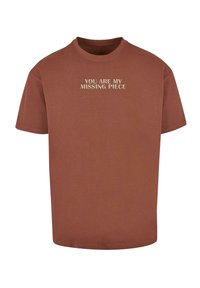 Brun bomulds T-shirt med korte ærmer og rund hals. På forsiden står der tekst "DU ER MIT MANGLENDE PUZZLESTYKKE" i en kontrasterende lys farve.