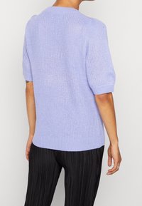 Person in einem helllila gestrickten kurzärmeligen Pullover mit gerippten Bündchen und Saum, kombiniert mit schwarzen vertikal gefalteten Hosen.