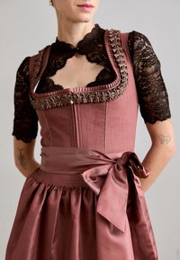 Femme portant une robe traditionnelle rose poussière avec des bordures brodées, un chemisier en dentelle noire et une ceinture en satin nouée en un nœud sur le côté.