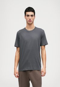 Γκρι βαμβακερό μπλουζάκι με κοντά μανίκια και στρογγυλή λαιμόκοψη, σε μοντέλο που φοράει καφέ παντελόνι. Απλός σχεδιασμός, χωρίς μοτίβα ή λεπτομέρειες.