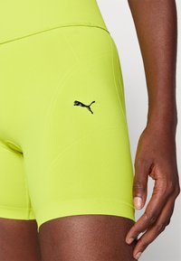 Zbliżenie na biodro i rękę osoby ubranej w jaskrawożółte spodenki sportowe Puma z logo na udzie na jednolitym tle.