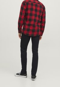 Man i röd och svart rutig skjorta, svarta slim fit byxor och svarta låga sneakers med vita sulor, vänd bort mot en neutral bakgrund.