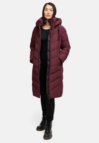 Cappotto piumino bordeaux con cappuccio, dotato di chiusura con zip, cuciture a zigzag e cordini regolabili, abbinato a pantaloni neri e stivali.
