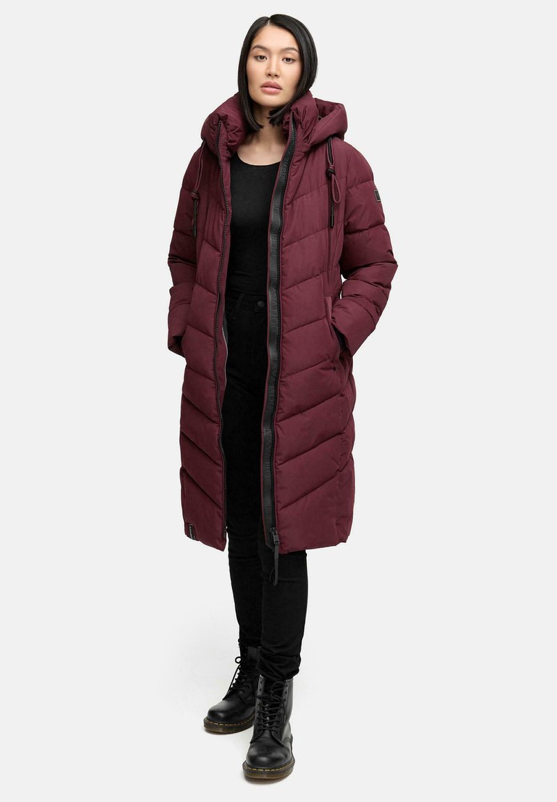 Cappotto piumino bordeaux con cappuccio, dotato di chiusura con zip, cuciture a zigzag e cordini regolabili, abbinato a pantaloni neri e stivali.