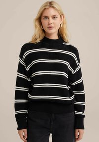 Svart stickad tröja med vita vågräta ränder, hög krage, lös passform och ribbade ärmslut. Texturen ser grov och varm ut.