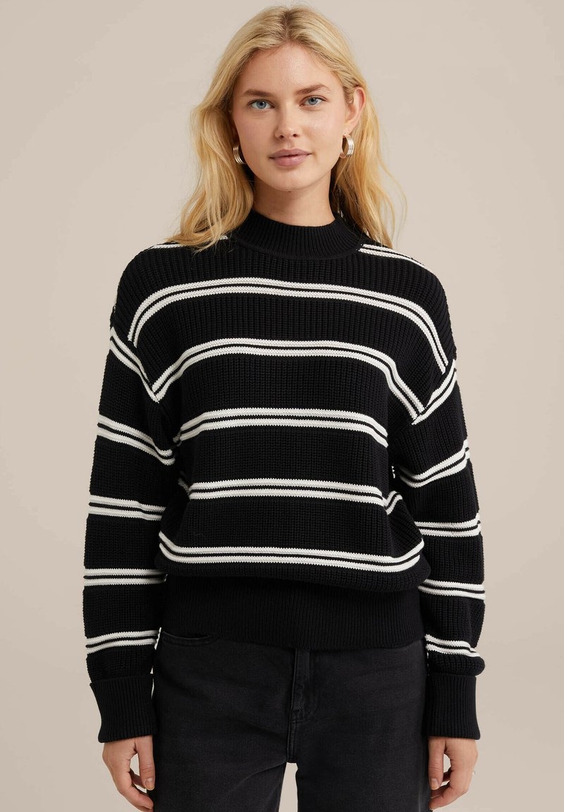 Svart stickad tröja med vita vågräta ränder, hög krage, lös passform och ribbade ärmslut. Texturen ser grov och varm ut.