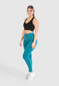 Sujetador deportivo negro de canalé y leggings de talle alto en color verde azulado, ambos hechos de tela elástica. Zapatillas blancas completan el atuendo atlético.
