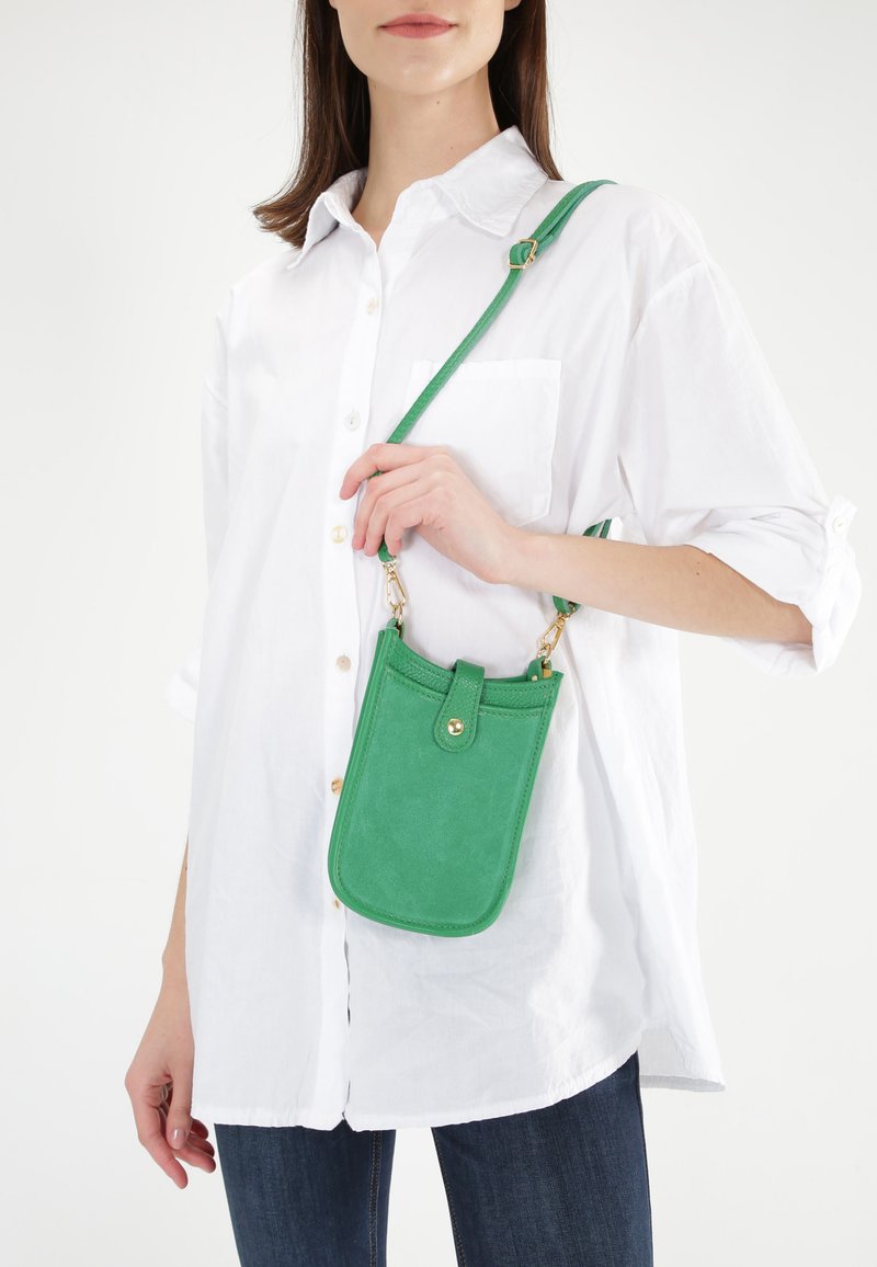 Grøn suede crossbody taske med klap, guld hardware og justerbar rem, båret af en person iført hvid skjorte og blå jeans.