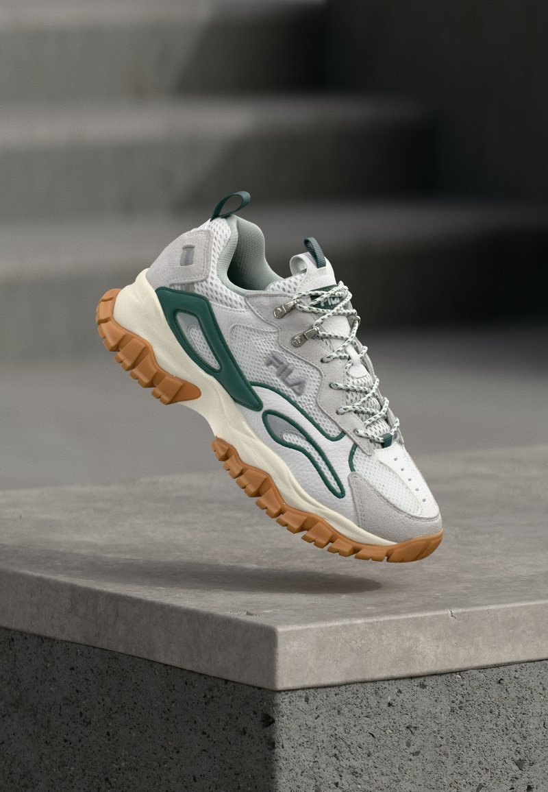 Witte en groene sneaker met een dikke bruine zool zwevend boven een betonnen platform met vervaagde traptreden op de achtergrond.