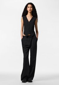 Sieviete ar gariem cirtainiem matiem, valkājot melnu, bezroku vestes un vaļīgas melnas svītrainas bikses, stāv ar rokām kabatās.