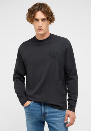 Schwarzes Langarmhemd mit Rundhalsausschnitt, das ein dezentes erhabenes Logo auf der Brust trägt. Getragen mit blauen Jeans, die eine lässige Passform zeigen.