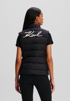 Schwarze Pufferweste mit hohem Kragen und horizontalen Nähten, mit dem weißen, gestickten "Karl"-Logo auf dem Rücken. Getragen über einem schwarzen Shirt.