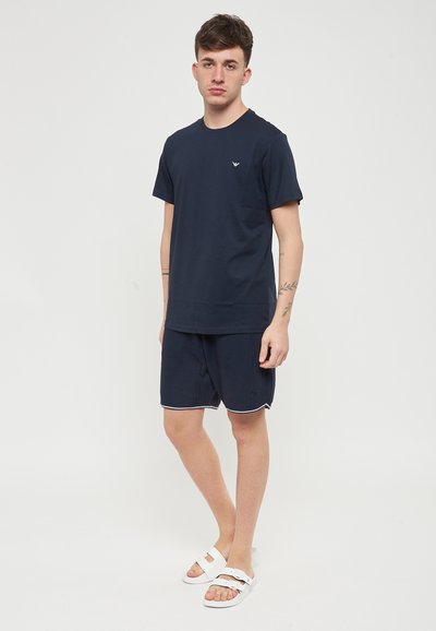 T-shirt blu navy con collo rotondo e un piccolo logo, abbinato a pantaloni corti blu navy con un design testurizzato e bordi bianchi, indossato con sandali bianchi.