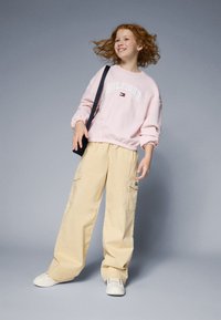 Sweatshirt rose clair avec le logo "HILFIGER", pantalon cargo beige et baskets blanches. Le mannequin pose avec un sac à bandoulière noir.