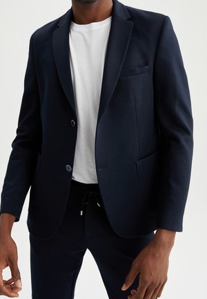 Homme portant un blazer bleu marine sur un t-shirt blanc avec un pantalon bleu marine assorti, debout devant un fond uni.