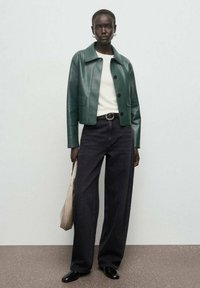 Chaqueta de cuero verde ajustada con cierre de botones, suéter blanco, jeans oscuros de pierna ancha y zapatos negros. Bolso beige con detalle texturizado.