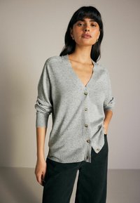 Lysegrå cardigan med V-hals, knaplukning fortil og ribbede manchetter. Har en plettet tekstur og en afslappet pasform, kombineret med mørke bukser.