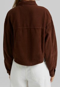 Hnědá kordurová krátká bunda s límcem, viditelnou vertikální žebrovanou texturou a zapínáním na knoflíky na manžetách, kombinovaná s bílými kalhotami.