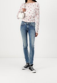 Mujer con una blusa de encaje floral de mangas largas, jeans ajustados azules, zapatillas de lona altas negras y sosteniendo un pequeño bolso blanco sobre el hombro.