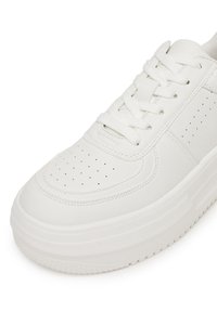 Prim-plan al unei tenisi platformă albe cu vârf din piele perforată, talpă groasă și șireturi albe legate într-o fundă.