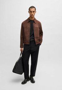 Giacca in suede marrone con tasche frontali, t-shirt nera, pantaloni neri e scarpe lucide nere; in mano una borsa nera. Sfondo neutro.