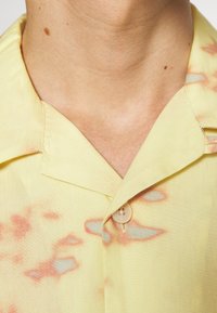 Lichtgele geknoopte blouse met abstracte roodachtige patronen en een zachte textuur. Korte mouwen en een standaard pasvorm.