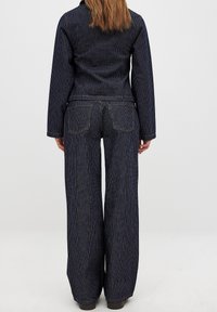 Completo in denim composto da un giubbotto a righe blu scuro e pantaloni larghi coordinati. Entrambi i pezzi presentano cuciture a contrasto e tasche posteriori.