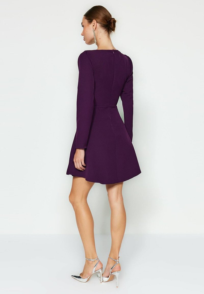 Robe longue violette à manches longues avec une taille cintrée et une jupe évasée. Dotée d'une fermeture éclair au dos. Portée avec des chaussures à talons hauts argentées.