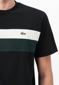 Schwarzes T-Shirt mit horizontalen Streifen in Creme und Dunkelgrün, mit einem kleinen grünen gestickten Krokodil-Logo auf der Brust. Kurze Ärmel.