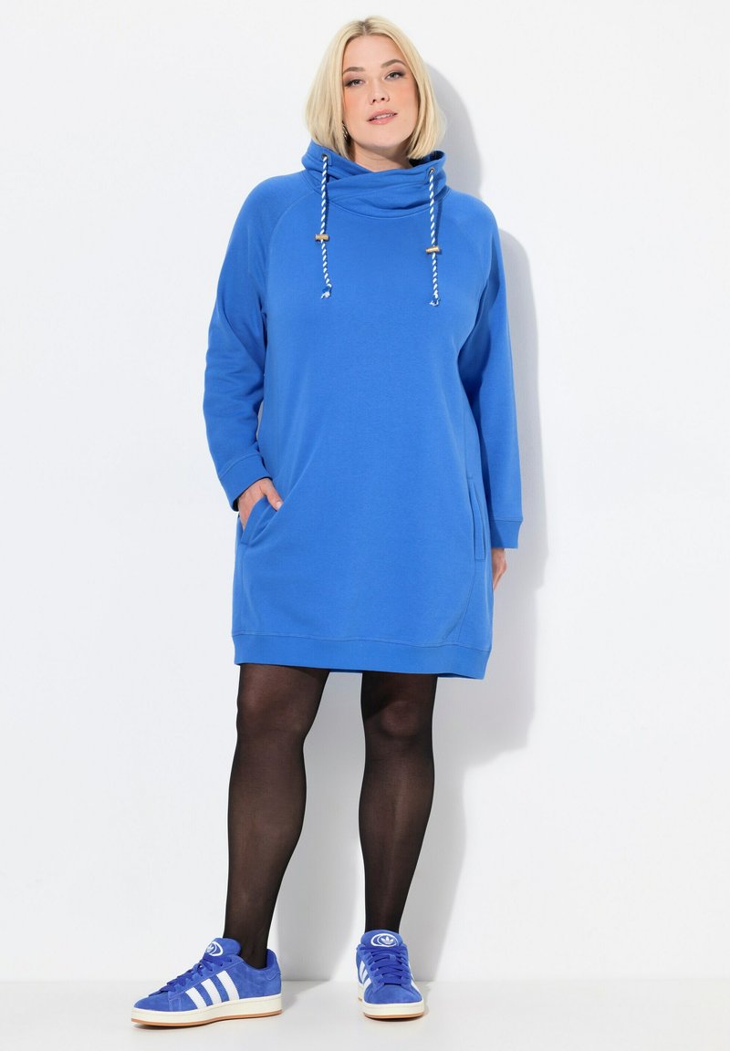 Kobaltblaues Kapuzenkleid mit Fronttaschen, Zugbändern und gerippten Bündchen. Getragen mit schwarzen Strumpfhosen und blauen Sneakers.