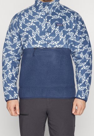 Mann trägt einen blau-weiß gemusterten Patagonia-Fleecepullover mit einer Reißverschlusstasche auf der Brust und einem Kragen mit Druckknöpfen.
