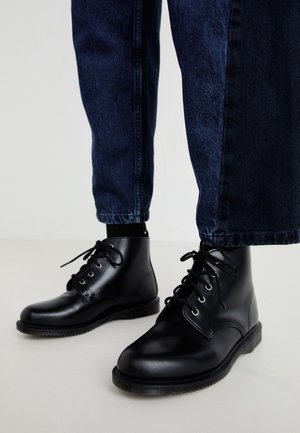 Bottes en cuir noir montant à la cheville avec bouts arrondis, lacets à l'avant et œillets métalliques, associées à un jean bleu foncé. Texture lisse et semelle robuste.