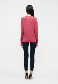 Κάρποι πουλόβερ μακρυμάνικο σε βαθυκόκκινο με στρογγυλή λαιμόκοψη και ριμπ μανσέτες, συνδυασμένο με σκούρο στενό τζιν και μαύρα ψηλοτάκουνα παπούτσια.