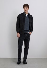 Man med svart mocka bomberjacka, mörkgrå zip-up tröja över vit skjorta, svarta jeans och svarta snörskor står mot en enkel vägg.