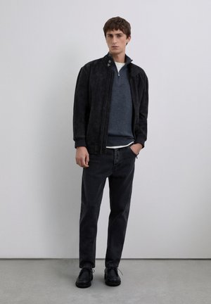 Man wearing black suede bomber jacket, dark gray zip-up sweater over white shirt, black jeans, and black lace-up shoes standing against plain wall.