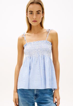 Frau trägt ärmelloses, blau-weiß gestreiftes, gerafftes Oberteil mit Bindeträgern und blaue Jeans, steht vor einem einfarbigen Hintergrund.