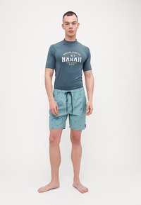 Tričko s krátkym rukávom v odtieni teal so bielym textom "Hawaii"; skombinované s teal plavkami s malými farebnými surfovými postavami.