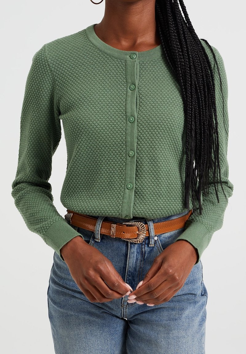 Green textured cardigan with a round neckline, front button closure, and long sleeves, paired with high-waisted denim and a brown belt.