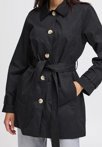 Čierny trench coat s opaskom a golierom, zlatými gombíkmi a prednými vreckami. Mäkko pôsobiaca textúra s hladkým povrchom.