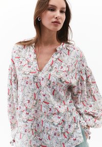 Blusa floreale con scollatura a V, maniche lunghe, tessuto leggero con motivi rosa e grigi, e dettagli arricciati alle spalle.